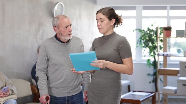 Positive polite young secretary or social worker talking to aged man visitor in reception area in background queue of people 