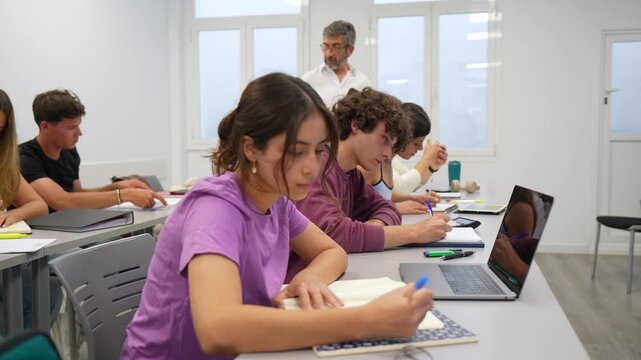 Focused young college students writing in their notebooks while sitting at desks in a modern classroom, with a male professor walking in the background and supervising their work during a lesson