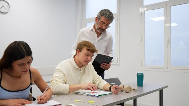 Mature male teacher helping a young male student during a lesson while his classmate is taking notes. Education, mentorship, and learning concept in a modern university or college classroom setting