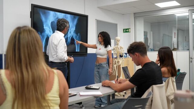Young female student pointing at a pelvic x ray on a large screen during an anatomy class, while the professor explains and classmates take notes at their desks in a modern university classroom