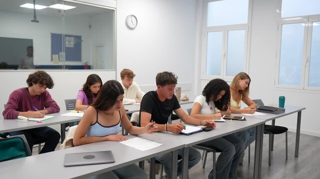 Diligent high school students taking a test in a classroom, where one male student raises his hand to ask a question and the mature professor approaches to provide assistance and clarification