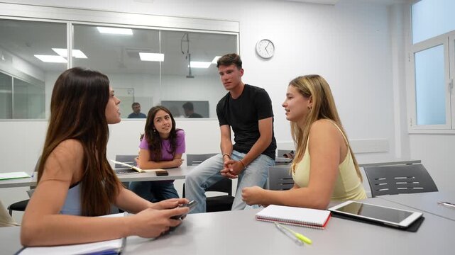 Group of four diverse young students studying together, actively discussing ideas and collaborating on a university assignment in a modern, bright classroom setting with notebooks and tablets