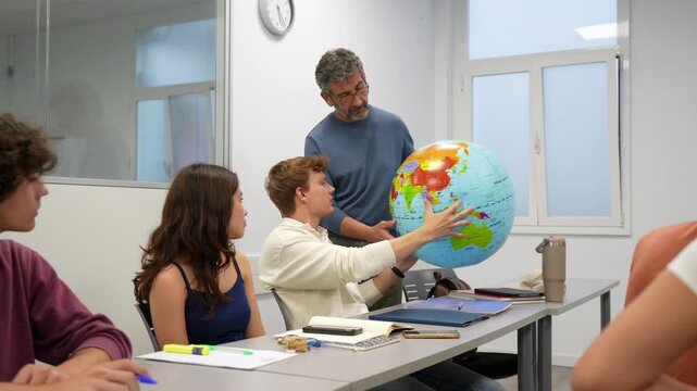 Mature male teacher with a group of diverse university students during a geography class, explaining continents on an inflatable globe while everyone listens attentively in the classroom