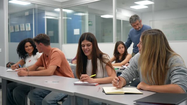 Diverse group of young college students laughing and talking in a modern classroom while taking notes, with their mature professor standing and supervising them in the background