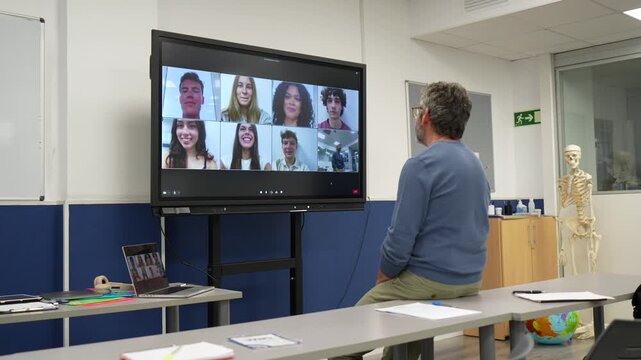 Middle aged male professor interacting with a group of students during a distance learning class, using a large interactive whiteboard for a video conference in a modern, empty classroom