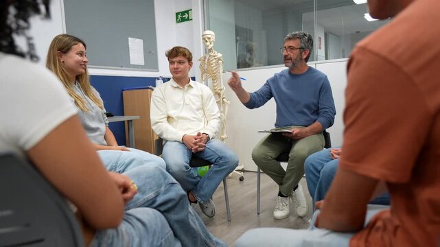 Mature male teacher holding a clipboard and gesturing while explaining a biology lesson to a group of university students sitting in a circle, with a human skeleton model in the background