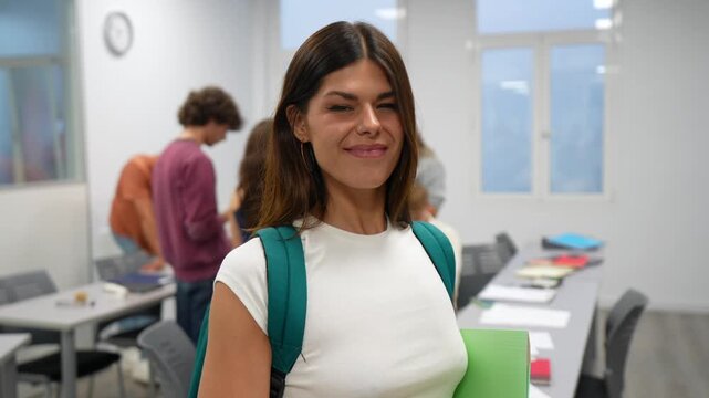 Cheerful female student with a backpack and folder winking and smiling at the camera, standing in a university classroom while other classmates are visible in the blurred background