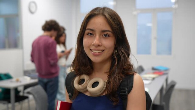 Portrait of a happy young hispanic female student holding a book and looking at the camera, standing in a bright classroom with other classmates interacting in the blurred background