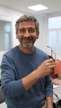 Middle aged university teacher taking off his glasses and smiling at the camera with a satisfied expression, while students are sitting at desks in the classroom during a lesson