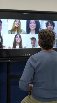 Male teacher sitting in front of a smart board during an online lesson, engaging with a grid view of his smiling students participating remotely from home through a video conference call