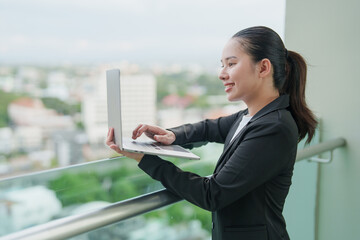 Fototapeta premium Asian businesswoman working remotely on a laptop, standing happily outdoors on a high-rise balcony, managing a global business with city views, using technology for a digital lifestyle
