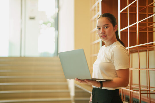 Young Asian woman holding an open laptop, looking at the camera, standing in a brightly lit modern interior with wooden steps and shelves, symbolizing connectivity