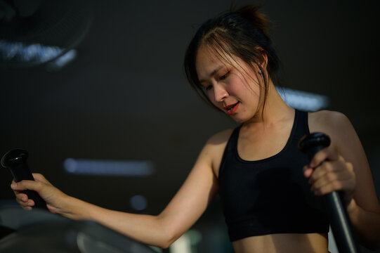A focused woman in a black sports bra exercises intensely on gym equipment in a dimly lit setting.