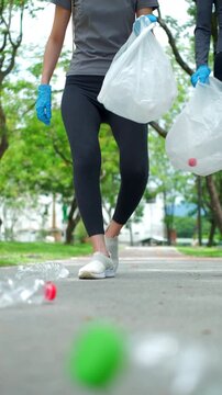 Two young Asian women volunteers in activewear picking up plastic bottles from a park road into trash bags. Concept of environmental conservation, community service, and eco-friendly.