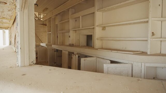 Empty white shelves line a faded kitchen space. Rustic cabinetry stands silent, showing years of neglect. Dust covers countertops and doors, giving a quiet, abandoned feel