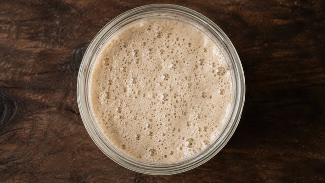 Yeast sourdough starter in a glass jar with a frothy surface, placed on a rustic wooden table, showcasing the fermentation process of bread dough rising