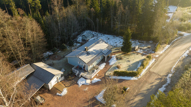 Aerial view of a detached house with a wooden deck surrounded by a dense forest and patches of snow on the ground next to a rural road in Uusimaa, Finland.