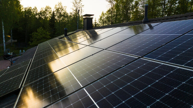 Aerial view of solar panels installed on a house roof reflecting the evening sun with lush green trees in the background in Uusimaa, Finland.