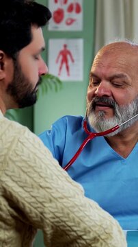 Indian male doctor treating patient during routine health checkup as experienced physician examines young man with stethoscope in modern Indian clinic OPD healthcare consultation visit