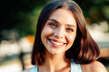 Fototapeta premium Smiling young woman portrait outdoors with natural light and green bokeh background, warm summer mood and friendly confident expression