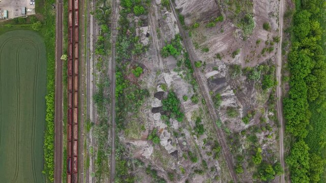 An aerial drone perspective captures a freight train on tracks, contrasting a lush green field with a stark industrial landscape in Hungary.