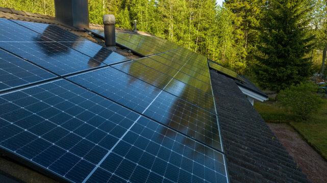 Aerial view of modern solar panels installed on a dark tiled roof surrounded by lush green forest in Uusimaa, Finland.