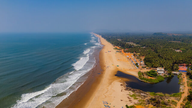 Aerial view of the long sandy coastline with waves crashing on the shore and lush palm trees bordering the beach in Gokarna, Karnataka, India.