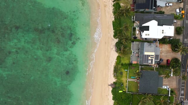 Luxury beachfront homes with solar-paneled rooftops and lush palms line a narrow white sand beach above stunning clear turquoise coral reef waters in Laie, Hawaii, USA. Aerial drone top down.