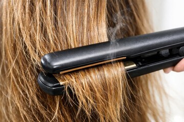 Naklejka premium A close-up of a hair straightener smoothing blonde hair, with visible steam rising as heat is applied.