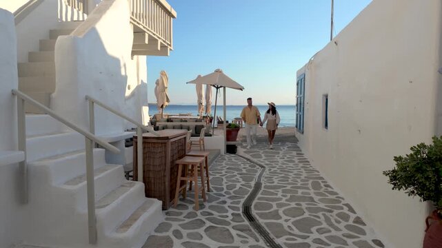 A diverse couple explores the delightful pathways lined with whitewashed buildings in Naousa, Paros, Greece. Experience the warm sunlight and charming atmosphere in this Cycladic gem.