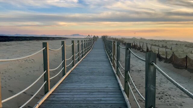 4K static shot of a wooden boardwalk at Labenne Oc&eacute;an, France, leading to the beach with the Pyrenees mountains visible in the distance at sunset.