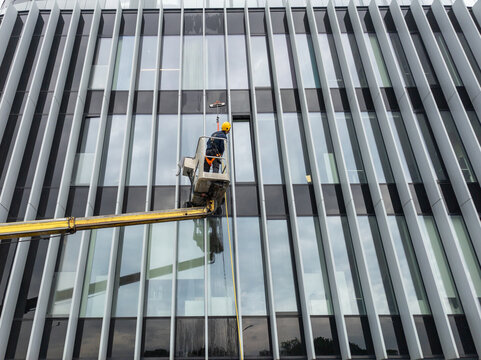 Professional window washer cleaning glass facade of modern office building using cherry picker crane, maintenance worker washing windows on high rise skyscraper