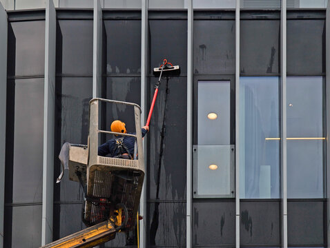 Professional window washer cleaning glass facade of modern office building using telescopic pole from a cherry picker platform, industrial maintenance service