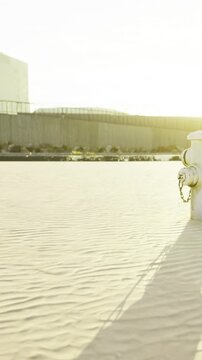 A water source is placed in a sandy area with a wall in the background. The sunlight creates long shadows. The scene shows an urban environment at the end of the day.