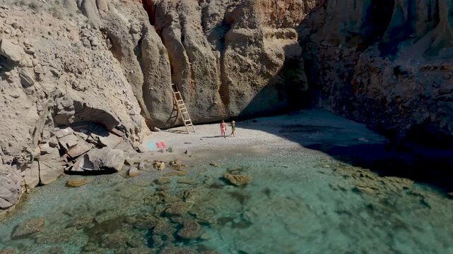 Explore the serene beauty of with a couple of men and women walking at the Tsigrado Beach, Milos, Greece, where turquoise waters meet dramatic cliffs. 