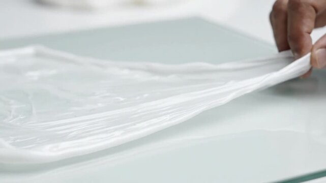 A hand slowly lifts a transparent, thin, and flexible sheet, showcasing its delicate texture on a light background, ideal for science and technology visuals