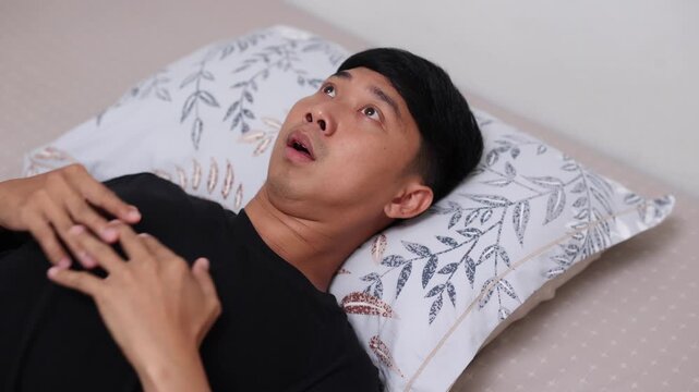 Asian man lying in bed suddenly wakes up feeling terrified, gasping for air and looking around in panic