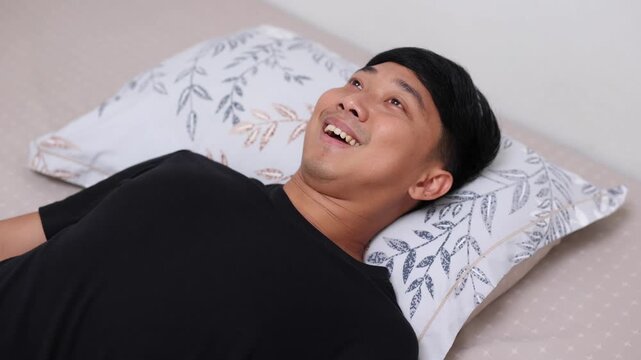 cheerful Asian man lies in bed, smiles warmly, pulls up a cozy patterned blanket, and closes his eyes to sleep