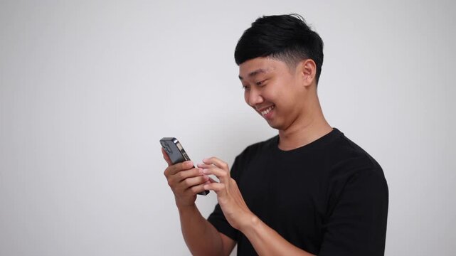 A cheerful Asian man in a black t-shirt scrolls on his mobile phone and smiles directly at the camera against a plain background