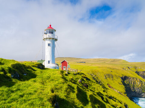 Akraberg Lighthouse on Suduroy Island, Faroe Islands. Sunny summer day landscape with green meadows, cliffs and ocean under a cloudy sky. Postcard background.