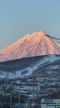 Majestic snow-covered volcano mountain under the first rays of sunrise, with dramatic light illuminating the peaks. A scenic road leads toward the mountain with a car in motion, creating a sense of jo