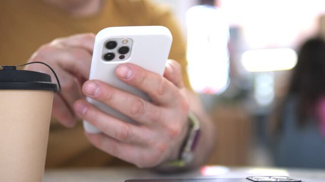 Person holds phone, taps screen. Coffee cup rests beside hand. Blurred background with warm lighting. Focus stays sharp on phone and fingers. No animals or plants visible