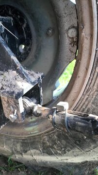 Close-up mechanical scene showing the rear section of a tractor with a visible wheel and metal frame. A male hand inserts a bolt into a hitch mounting point and secures it using a fastening clamp