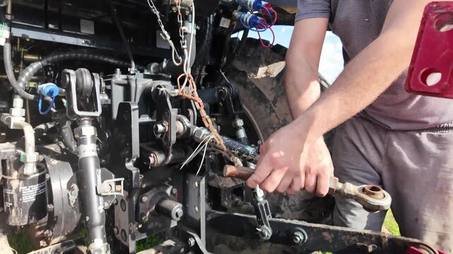 Rear view of a modern tractor showing mechanical components of the hydraulic linkage system. A male operator removes a locking pin from the rear hitch connection, releasing the attachment point. The