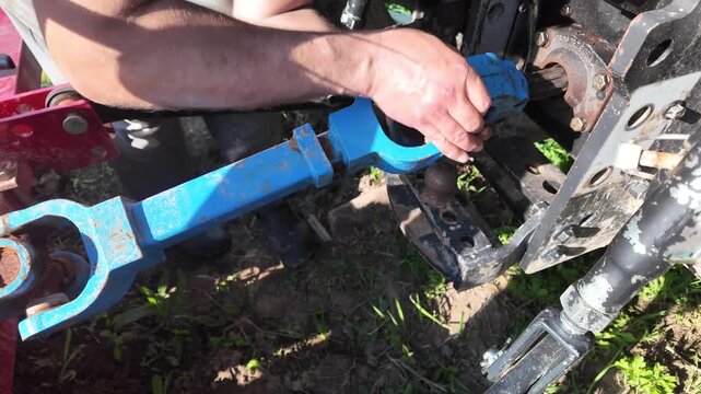 Close-up footage of a mans hands lifting and attaching a PTO drive shaft (cardan shaft) from the ground and connecting it to a tractor. The operator aligns the coupling, pushes it into place, and
