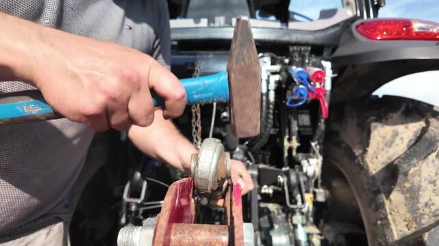 Slow motion close-up of a male operator inserting a heavy tractor linkage arm into rear mounting brackets of a rotary implement. The metal component enters the tight-fitting eyelets with visible