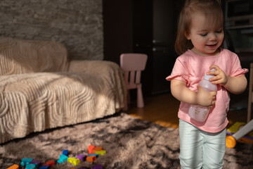 Naklejka premium smiling toddler in a pink shirt holding a floral baby bottle, standing on a sunlit plush brown rug in a cozy living room scattered with colorful toy blocks beside a beige sofa and pink chair