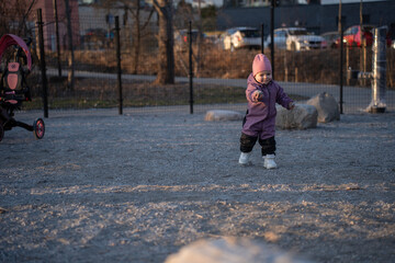 Fototapeta premium toddler in a purple snowsuit and pink beanie walking across a gravel urban park at golden hour, reaching forward near rocks and a stroller, warm light and soft shadows in a candid playful moment