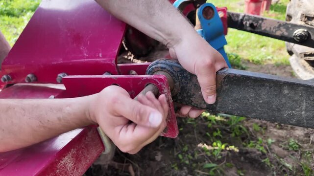 Close-up shot of male hands manually aligning and inserting a locking pin into a tractor rear hitch connection. The hydraulic linkage from the tractor connects into a bracket system on a rotary