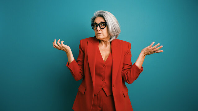 Senior businesswoman with grey hair and glasses wearing a red suit, shrugging her shoulders, portraying confusion on a teal background